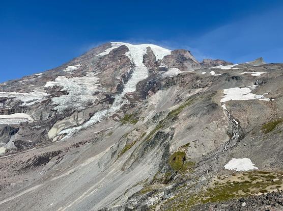 A mountainous Mt Rainier landscape featuring the Nisqually glacier, surrounded by rocky terrain under a clear blue sky. The mountain has patches of snow & glaciers tumbling down its steep slopes.