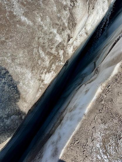 A narrow, dark water channel flows between two walls of glacial snow known as a crevasse. The crack appears quite deep, and its too dark to know how deep it is!