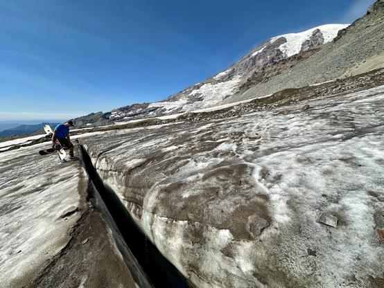 A snowboarder looks down a large crack in a glacier. A massive Mt Rainier can be seen in the background of a clear blue sky.