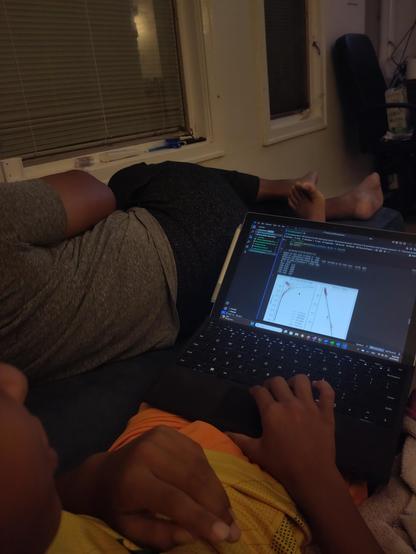 Photo taken over the shoulder of a child working on a tablet in a dimly lit room. His cheek and nose can be seen in the lower left corner, with the tablet on his lap. He has a yellow shirt and orange shorts, and his right hand is on the mouse pad while his left hand is across his chest. The computer screen shows some code and a graph. Windows on a wall and another person sprawled on the couch are visible in the background.