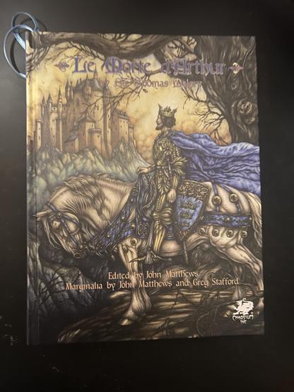 Le Morte D’Arthur by Sir Thomas Malory.

Annotated by John Matthews and Greg Stafford. 

King Arthur wearing a golden crown and blue cape. He rides a white horse and gazes at a castle in the distance.