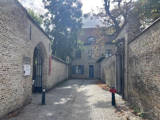 Photo of the entrance to the Groeningemuseum in Bruges, on a late summer's day, moving towards autumn. The sun is shining, the trees are still green, but a few leaves on the ground signal the transition. The entrance consists of a black forged iron gate through a brick wall.