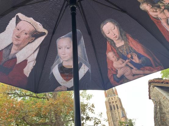 An umbrella from the museums of Bruges, the exterior is navy blue, the interior features representations of women from paintings by Memling, Van Eyck, and others. The umbrella is photographed open, with a building in Bruges visible in the background.