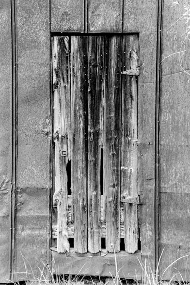 A dilapidated wooden door to a utility shed.  The shed is clad with galvanized metal siding.  It's a black and white film image, the contrast is relatively high and the grain present, but in my opinion pleasantly so.