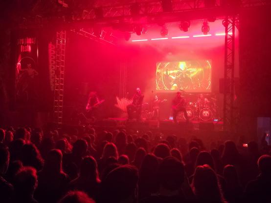 A metal band performs on stage, a good part of the audience can be seen in front of the stage. In the background is an animated pentagram.