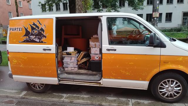 Das Bild zeigt ein orangefarbenes und weißes Van, das an einer Straße geparkt ist. Die Tür des Vans ist geöffnet, und man sieht den Innenraum, der mit verschiedenen Kartons und Behältern gefüllt ist. Auf der Seite des Vans ist ein schwarzes Logo mit einem Piratenmotiv und dem Wort "PARTEI" zu sehen. Im Hintergrund sind Gebäude und Bäume zu erkennen. Der Van scheint für den Transport von Waren oder Materialien zu verwendet werden.