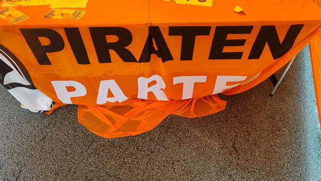 Das Bild zeigt eine Tischdecke in leuchtendem Orange, auf der in großen, schwarzen und weißen Buchstaben "PIRATEN PARTEI" steht. Die Tischdecke ist an einem Tisch befestigt, der auf einem grauen Teppichboden steht. Auf der Tischdecke liegen einige gelbe Broschüren oder Flyer. Die Farbe Orange ist charakteristisch für die Piratenpartei, eine politische Partei in Deutschland. Die Tischdecke ist eindeutig mit der Partei in Verbindung gebracht und dient als visuelles Symbol für die Partei.