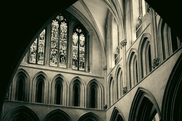 Interior of a gothic cathedral: looking slightly upwards from underneath an arch to the ceiling towards a row of arched openings and a big stained-glass window.