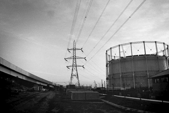 Black & white image of an urban scene, with an electricity pylon and a gas-holder.