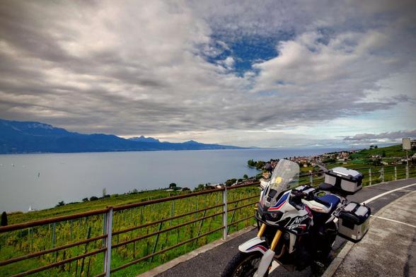 A motorcycle parked on a roadside viewpoint overlooking Lake Geneva, with vineyards in the foreground, villages along the shore, and mountains in the distance under a dramatic cloudy sky.