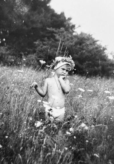 Schwarz Weiß Foto eines 2 Jahre alten Kindes auf einer Blumenwiese mit Bäumen und blauem Himmel im Hintergrund. 
Das Kind hat den Kopf nach links geneigt und stützt das Kinn auf die linke Hand. In der rechten Hand hält es eine Blume