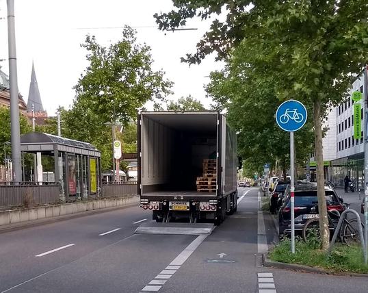 LKW parkt, mit abgesenkter Laderampe, links vom benutzungspflichtigen Radfahrstreifen, am rechten Rand der rechten von zwei Autofahrspuren.