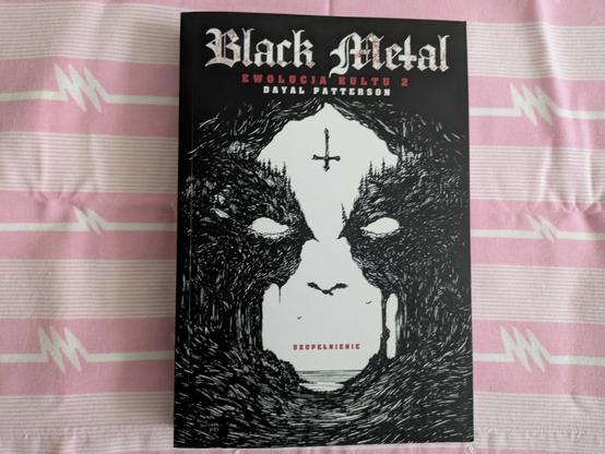 Okładka książki "Black Metal Ewolucja kultu 2. Uzupełnienie" Dayala Pattersona na tle różowej pościeli, tak dla żartu. Na okładce jest czarna maź, korzenie, które układają się w corpsepaintową twarz z wielkim ptakiem i odwróconym krzyżem na środku.