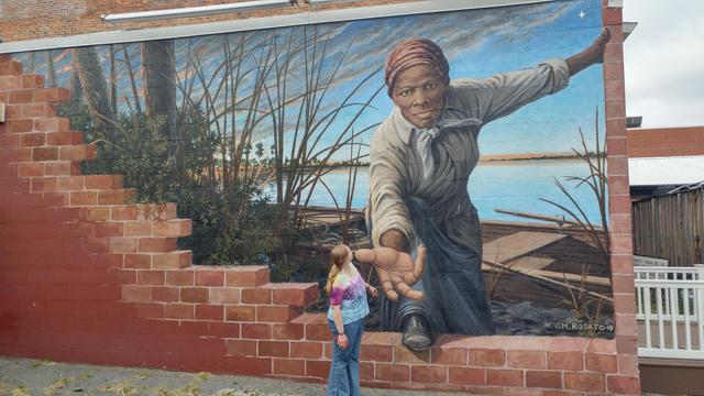 A woman looking up at a mural depicting Harriet Tubman reaching through a broken brick wall to help a runaway person.