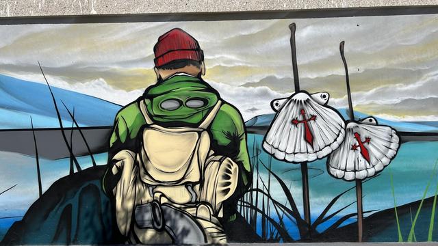 A colorful mural featuring a person in a green jacket and red beanie with their back to the viewer. They are surrounded by stylized seashells with red designs, against a backdrop of blue waters and a cloudy sky.