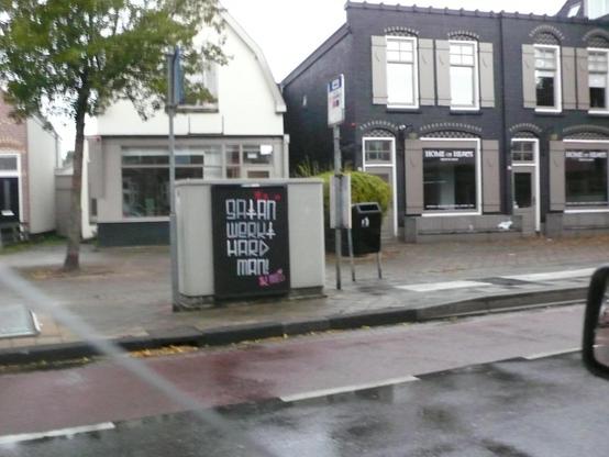 regenachtige straat met fietsstrook, rijtje panden, links een boom en bushalte bord. Straatkastje met zwart bord met witte letters. rechts boven wat rood en onderste letters in roze