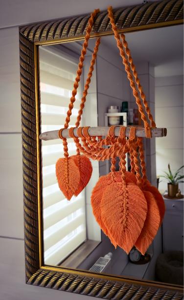 „Makramee-Wandbehang aus kräftig orangem Garn, in Form mehrerer Blätter, die an einem weißen Holzstab mit langen Fäden geknüpft sind
