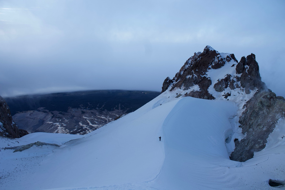 A lone climber traverses a snowy mountain slope, surrounded by rocky peaks and low-hanging clouds in a serene alpine landscape.