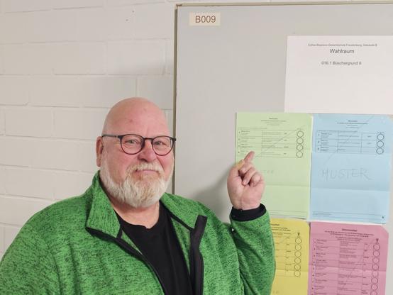 Das Bild zeigt einen Mann mit einem weißen Bart und einer Brille, der vor einer weißen Tafel steht. Er trägt eine grüne Jacke über einem schwarzen Shirt. Der Mann zeigt mit dem Zeigefinger auf ein Formular, das an der Tafel befestigt ist. Die Tafel ist mit verschiedenen Formularen in verschiedenen Farben versehen, darunter ein Formular mit der Aufschrift "Wahlraum" und ein Formular mit der Aufschrift "MUSTER". Die Tafel ist mit dem Bezeichner "B009" gekennzeichnet. Die Wand hinter der Tafel ist weiß und besteht aus Ziegelsteinen.

Bereitgestellt von @altbot, privat und lokal generiert mit Ovis2-8B