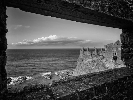 A black and white photograph of the Dunluce Castle ruins on a cliff edge overlooking the sea. The image is framed by a stone window in the foreground, creating a perspective as if looking out from inside the castle. The sea is calm, with a few rocks visible below the cliffs. The sky is partially cloudy.