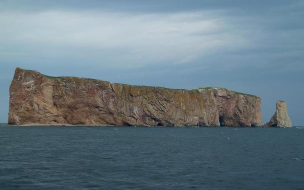Photograph of a large rock formation rising from the water, characterized by steep brown, red and ochre cliffs and a flat top, with greenery visible near the edge. A smaller rocky outcrop is situated to the right of the main formation, under a cloudy sky. Just barely visible from this angle is a hole in the rock, giving its name of "Rocher Percé".
