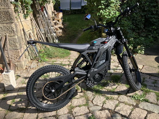 Schwarzes Elektro-Crossmotorrad, im Hintergrund eine Steinwand mit Gartengeräten und ein Trampelpfad