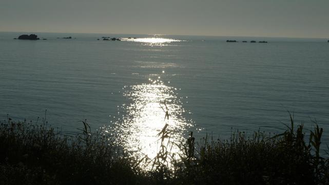 Gegenlichtaufnahme vom Meer. Auf dem Wasser glitzert es dort wo sich die tiefstehende Sonne darin spiegelt. Im Vordergrund ist Dünengras. In der Ferne sind einige Felsinseln zu sehen.