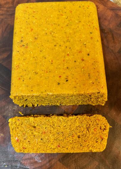 A block of a yellowish-green solid on a cutting board with one slice cut from the end. The texture appears to be like a fine-grained bread or cake. The added herbs and spices show as a few scattered dark specks.