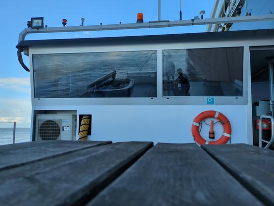 Schiffsbrücke von außen. In der Spiegelung der Scheiben sieht man ein Beiboot und eine Bank auf der ich sitze