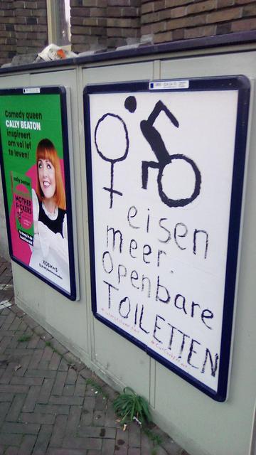 straatkastje aan bakstenenmuur met twee reclameborden. De voorste is bewerkt in een witte met in zwart, zie caption
De achterste is in groen met een persoon met rood haar