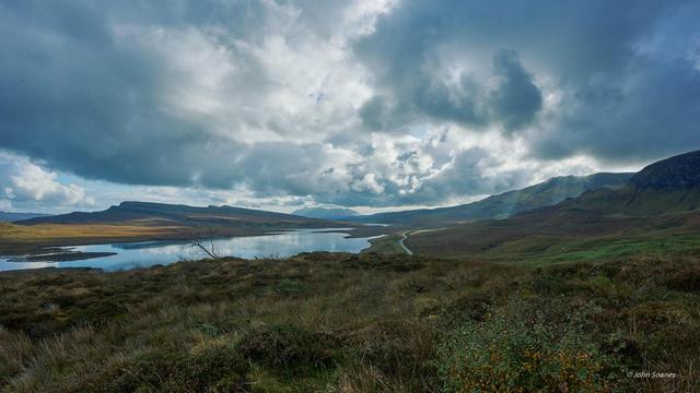 Dramatic skies over Loch Leathan.  The reflections from the clouds can be seen in the still waters of the loch.  There are sunbeams flowing down to the right of the road that winds through the back of the frame.  There are golden patches in the field illuminated by the sun around the rear and left side of the lake.