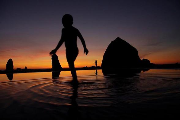 Photo of a small boy, silhouetted in the sunset, as he runs past a large rock formation in the distance, causing him to look like a giant. Haystack Rock on Cannon Beach in Oregon