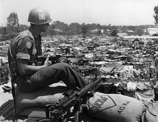 A Swedish peacekeeping soldier in the Congo, sitting on a tank, overlooking a vast refugee camp. The UN deployed troops from a variety of nations during ONUC.