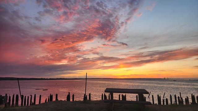 A color photo of a vivid sunset over Youngs Bay in Astoria, Oregon. In the foreground is a rough bench; behind it are pilings and two taller poles sticking up at the edge of the water. They are all black silhouettes. The water is illuminated by the sunset. The sky is orange yellow pink and purple.