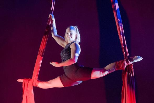 Sirkustaiteilija kiepauttanut jalkansa punaisten verhojen ympärille. Jalat ovat spagatissa pimeän teltan yläosassa. Taiteilijan katse suoraan kohti kameraa ja vain toinen käsi on kiinni verhossa.