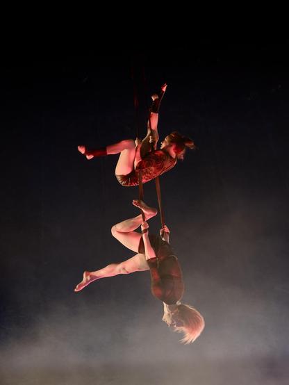 Kaksi sirkustaiteilijaa roikkuvat samoissa liinoissa. Toinen on pää alaspäin ja toinen hänen yläpuolellaan vaihtamassa asenota toisen jalan osoittaessa suoraan ylös. Kuvan alaossa muuta kuvaa valoisampaa savua.