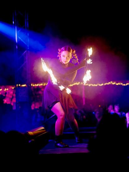 Lavalla olevan tulitaiteilijan käsissä on palavat esineet. Taiteilijan katse on suoraan kohti kameraa. Esiintymislavan värikkäät valot ovat värjänneet taiteilijan mustan asun ja kasvot pinkin sävyiseksi. Taustalla näkyy yleisöä epärerävänä.