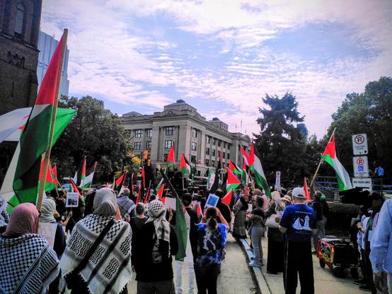 Photo of the crowd at a protest against the ongoing genocide in Palestine in London Ontario.There are a number of Palestinian flags visible and several people are wearing kufiyas. I took the photo from the back so that people's faces are not visible. The slightly cloudy blue sky is visible above.