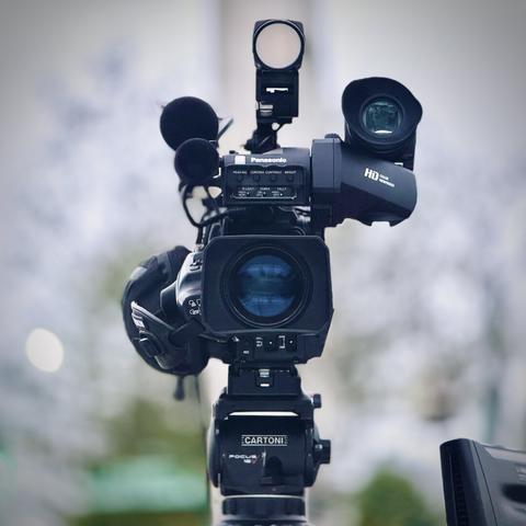 A black Panasonic electronic news-gathering (ENG) camera points straight ahead, resting on a black Cartoni video tripod under a cloudy sky.