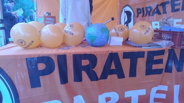 Das Bild zeigt eine Tischausstattung mit einer orangefarbenen Tischdecke, die mit großen schwarzen Buchstaben "PIRATEN" beschriftet ist. Auf dem Tisch sind mehrere gelbe Luftballons mit dem Wort "PIRATEN" und einem Logo platziert. In der Mitte des Tisches befindet sich ein Globus mit einem Metallstab. Im Hintergrund ist ein Banner mit dem Wort "PIRATEN" und einem Symbol zu sehen. Die Tischdekoration und die Ballons deuten auf eine Veranstaltung oder einen Stand der Piratenpartei hin.