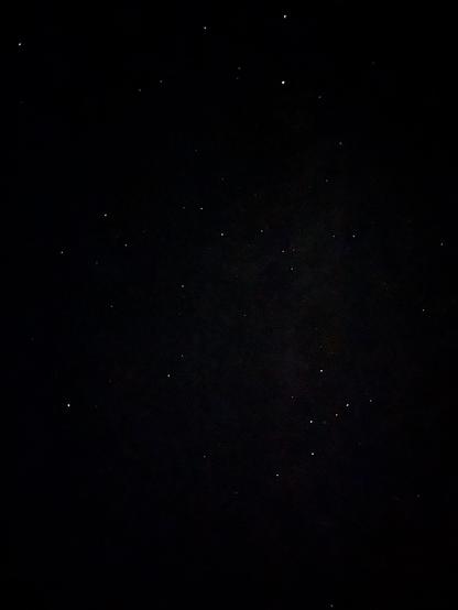 A dark night sky scattered with numerous small, bright stars. The majority of the stars appear white, with a few exhibiting a slight yellow or blue hue. The background is mostly black, indicating low light conditions typical of nighttime sky observations.