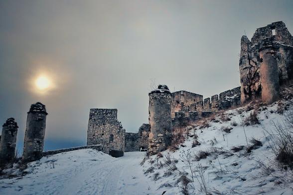 A wide-angle, low-shot photograph of ancient castle ruins on a snow-covered hillside. The sun is a bright, diffused orb in the grey sky. The stone walls and towers are a mix of large blocks and smaller, broken pieces, partially covered in snow. A path or road is visible, winding up the snowy hill towards the ruins.