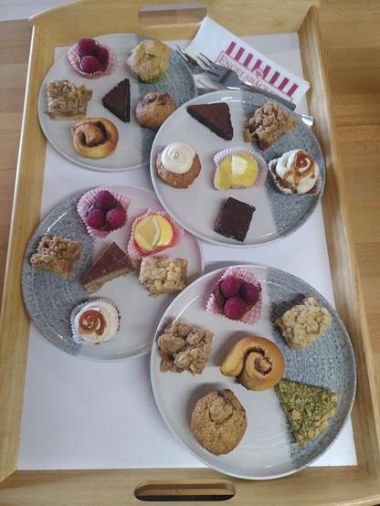 Auf einem Tablett mit hellem Holzrand befinden sich vier Kuchenteller mit je sechs kleinen Köstlichkeiten darauf. Das ganze nenne ich Probierteller. Mit dabei sind z. B. Cheesecakes mit Zitrone, Zimtschnecken, Bananenmuffins, Schokomoussetarte mit Himbeeren und Banoffee Pies (Banane-Karamell-Törtchen mit Sahnehaube).