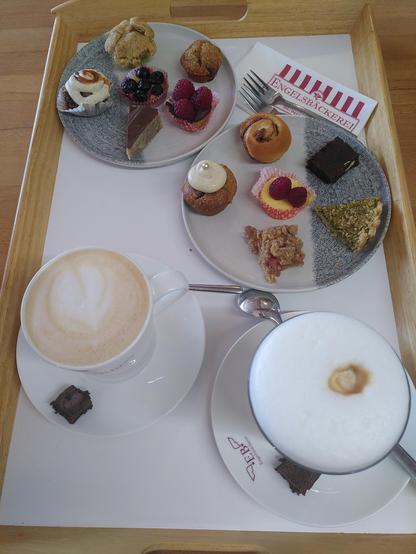 Auf einem weißen Tablett mit hellem Holzrand stehen vorne zwei Heißgetränke auf Untertassen (links ein großer Cappuccino, rechts ein Latte Macchiato) und dahinter zwei Probierteller mit je sechs kleinen Köstlichkeiten: Minitörtchen, Mini-Zimtschnecken, Mini-Apfelkuchen, Mini-Cheesecakes und anderes, insgesamt zwölf verschiedene Sorten.