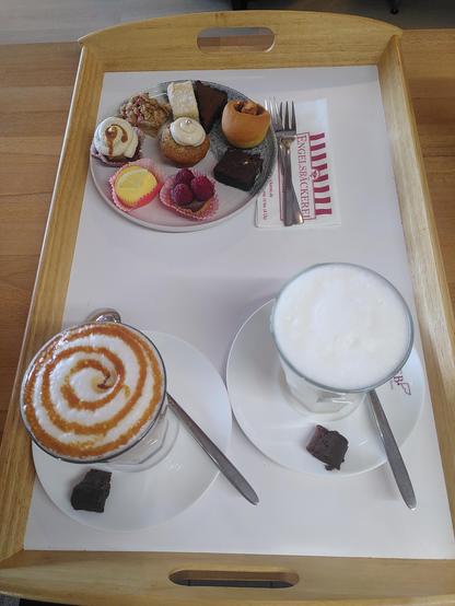 Auf einem weißen Tablett mit hellem Holzrand befinden sich vorne im Bild zwei Heißgetränke (links ein Caramel Macchiato (mit meinem selbstgemachten Karamell), rechts eine Honigmilch). Dahinter steht ein Probierteller mit neun kleinen Köstlichkeiten darauf (im Uhrzeigersinn): ein kleines Rechteck Shortbread mit Tonkabohne, ein klassischer Brownie in Dreiecksform, eine Zimtschnecke, ein Quadrat Espressobrownie, ein Stück Schokomoussetarte mit Himbeeren, ein Zitronen-Cheesecake, ein Banoffee Pie, ein Stück Apfel-Karamell-Kuchen und in der Mitte ein Bananencupcake mit Zimtfrischkäsecrème oben drauf.