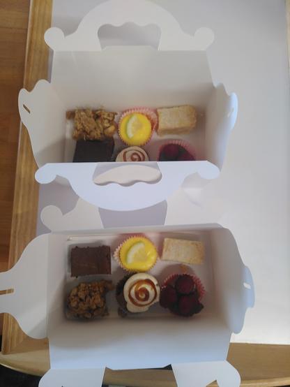 Zwei Probierteller mit je sechs kleinen Köstlichkeiten zum mitnehmen in je einer weißen Kartonage. Mit dabei sind ein klassischer Brownie, ein Zitronen-Cheesecake, ein Stück Shortbread mit Tonkabohne, ein Banoffee Pie, ein Stück Apfel-Karamell-Kuchem und ein Schokomoussetörtchen mit Himbeeren.