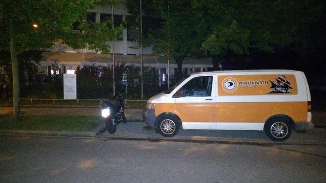 Das Bild zeigt eine nächtliche Szene an einer Straße. Auf der rechten Seite steht ein weiß-orangefarbener Van mit der Aufschrift "PIRATENPARTEI" und einem Piratenlogo auf der Seite. Der Van ist geparkt und hat seine Tür geöffnet. Links neben dem Van steht ein Motorrad mit einem Fahrer, der eine schwarze Motorradkleidung trägt und eine Laterne hält, die das Licht auf den Boden wirft. Im Hintergrund ist ein Gebäude mit Beleuchtung zu sehen, umgeben von Bäumen und Sträuchern. Die Straße ist beleuchtet, und die Umgebung wirkt ruhig.