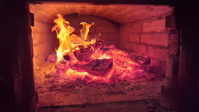 Burning wood in the combustion chamber of a traditional brick oven with a flat vault. Flames and decomposing wood are visible through the open iron oven door.