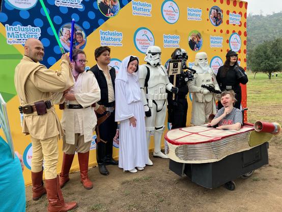 Star Wars cosplayers standing in a row, with a young man in a wheelchair landspeeder costume parked in front of them. Outdoors, in front of a backdrop. All are smiling.