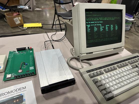 Photograph of a Wyse-30 terminal with a monochrome green monitor displaying various commands from a BBS. To the left of the terminal is a Hayes modem. Next to the modem is a sparsely populated circuit board.